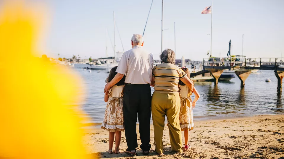 Photo of an elderly man and woman with there arms around two female children. They are standing on a beach looking at a harbour in the background.