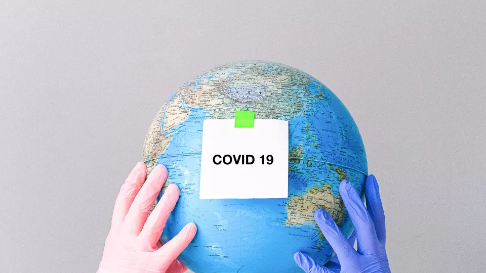 Two pair of hands in colourful gloves holding a small globe. There is a sign on the globe saying Covid-19.