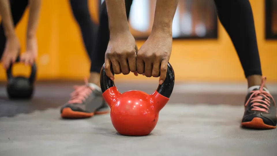 A kettlebell standing on the ground, two hands holding on to it and feet in the background