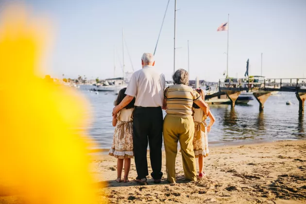 Photo of an elderly man and woman with there arms around two female children. They are standing on a beach looking at a harbour in the background.