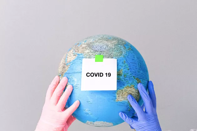 Two pair of hands in colourful gloves holding a small globe. There is a sign on the globe saying Covid-19.