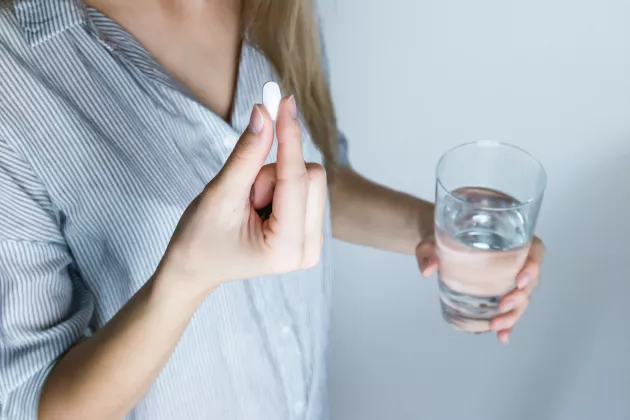 A close up picture of a person holding a white pill in one hand and a glas of water in the other. 