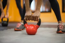 A kettlebell standing on the ground, two hands holding on to it and feet in the background