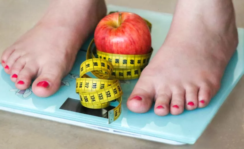 A photo of two feet on a blue digital scale. In between the feet on the scale is a red apple and yellow measuring tape.