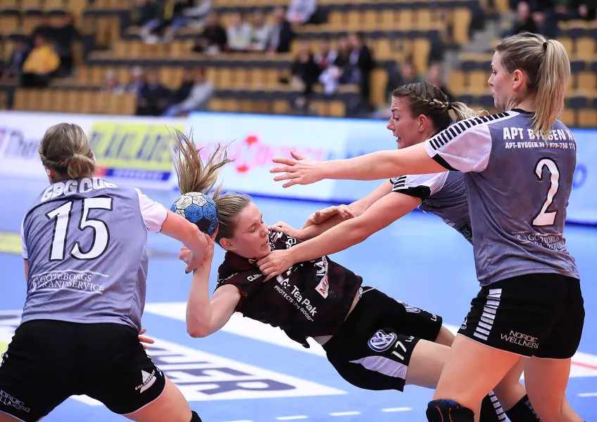 A photo of four female handball players playing handball. Three of the girls are in purple vests and are tackling a girl in a black t-shirt holding the ball.