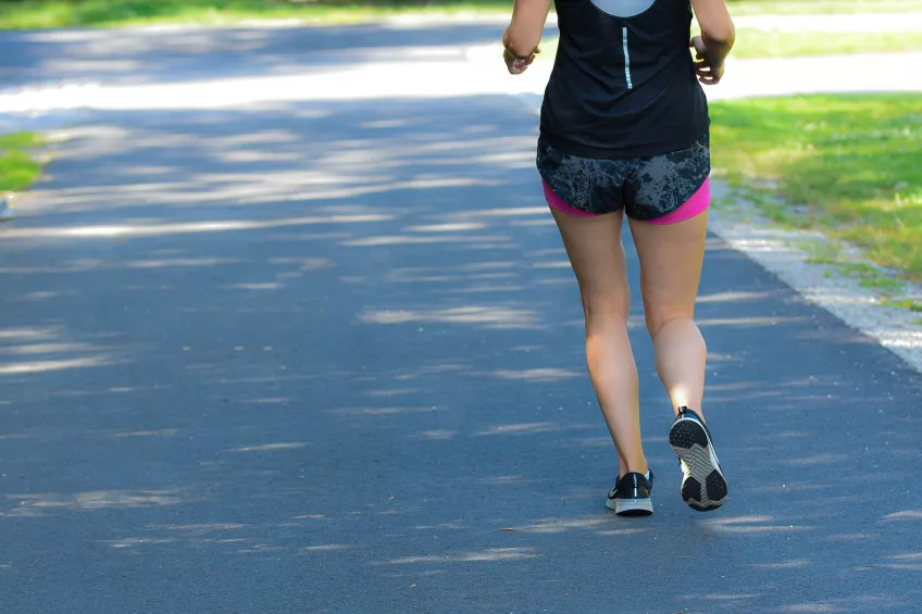A photo of a woman running on a concrete footpath. The woman is wearing a black tank top and black and pink shorts. The photo is taken from behind.