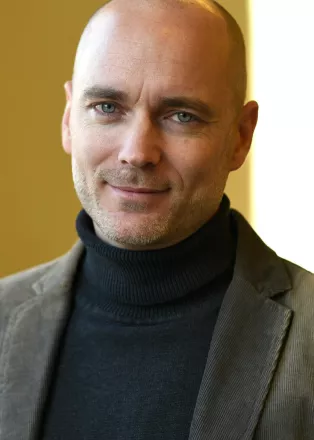 A portrait photo of Martin Englund. Martin is wearing a black turtleneck and a grey blazer and is smiling in the photo.