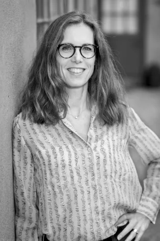A portrait photo of Eva Ageberg in black and white. Eva has long brown hair and is wearing round glasses and a striped blouse.
